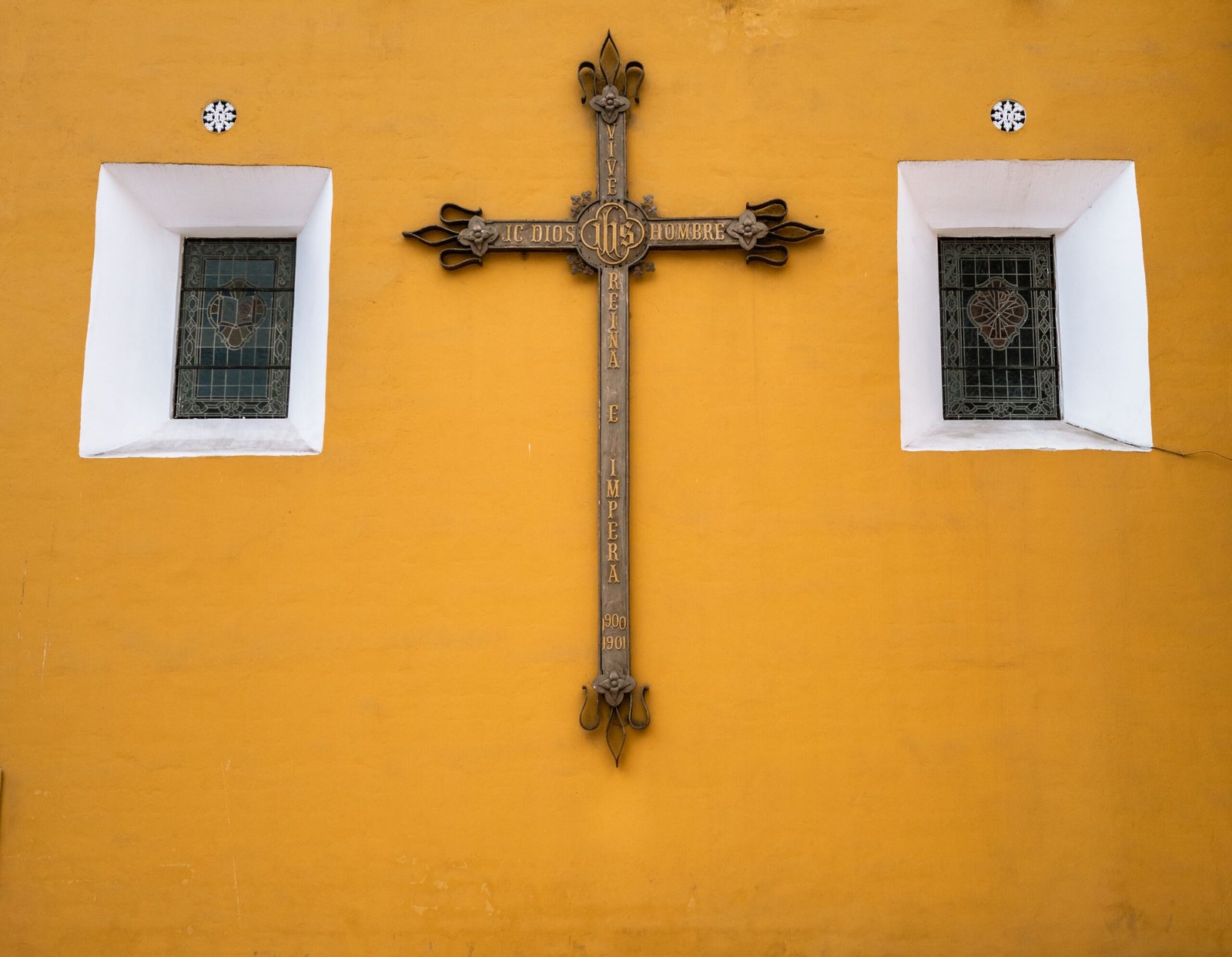 An ornate cross with religious text mounted on a vibrant yellow wall, flanked by two stained glass windows.